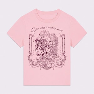 May include: Pink t-shirt with a graphic of two figures embracing, surrounded by ornate scrollwork. The text "Once upon a broken heart" is arched above the figures. The design is in a darker shade of pink.
