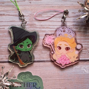Puede incluir: Dos colgantes acrílicos con personajes de dibujos animados. Uno representa a una bruja de piel verde con sombrero y vestido negro, sosteniendo una escoba. El otro muestra a una princesa con vestido y corona rosa, sosteniendo una varita. Ambos tienen llaveros.