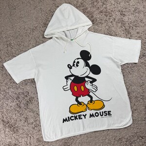 Może przedstawiać: Biała koszulka z kapturem z grafiką Myszki Miki. Postać z kresk&oacute;wki jest w kolorze czarnym, czerwonym, ż&oacute;łtym i białym. Słowa "Mickey Mouse" są wydrukowane na czarno pod obrazem.