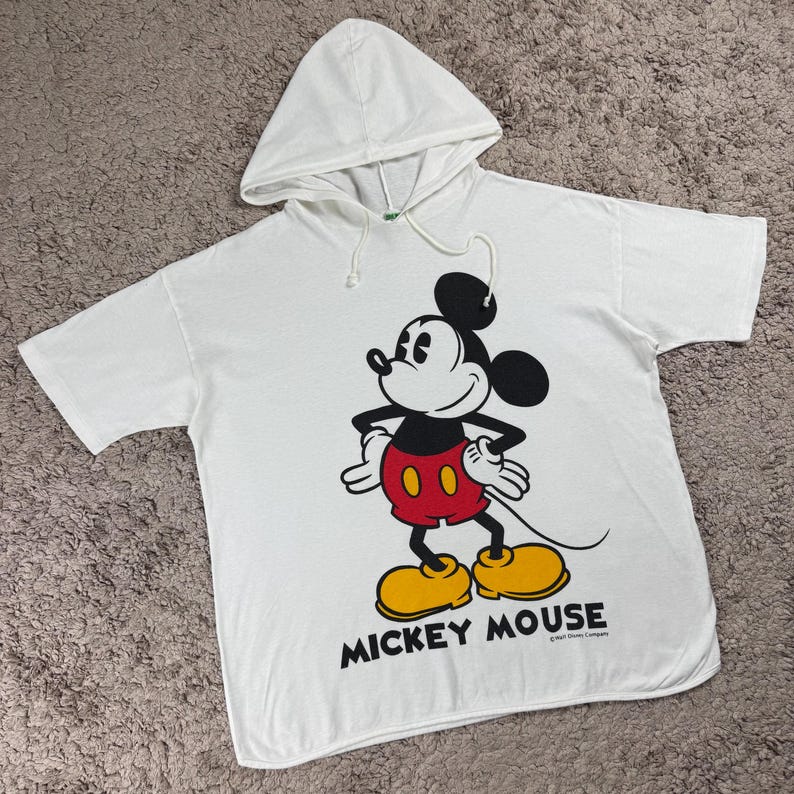 Może przedstawiać: Biała koszulka z kapturem z grafiką Myszki Miki. Postać z kresk&oacute;wki jest w kolorze czarnym, czerwonym, ż&oacute;łtym i białym. Słowa "Mickey Mouse" są wydrukowane na czarno pod obrazem.