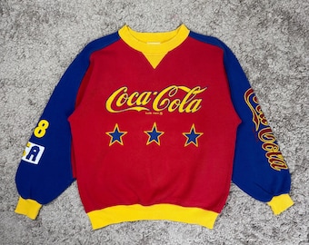 May include: A vintage Coca-Cola sweatshirt in red with blue sleeves and yellow trim. The front features the Coca-Cola logo in yellow, three yellow stars, and the text "8 A" on the left sleeve. The right sleeve has the Coca-Cola logo.