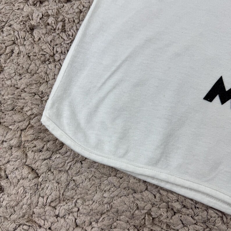 Może przedstawiać: Biały t-shirt z zakrzywionym dołem. Tkanina jest gładka i wydaje się być lekkim materiałem. Czarna litera jest widoczna na koszulce. Koszulka leży na teksturowanej, jasnobrązowej powierzchni.