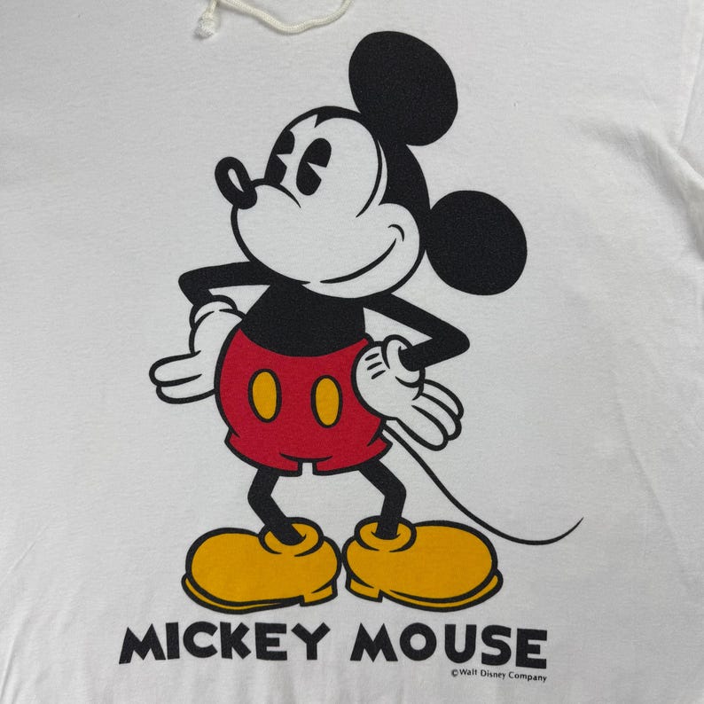 Może przedstawiać: Biała koszulka z grafiką Myszki Miki. Postać z kresk&oacute;wki jest w kolorze czarnym, czerwonym, ż&oacute;łtym i białym, z napisem "MICKEY MOUSE" wydrukowanym poniżej. Widoczny jest r&oacute;wnież copyright Walt Disney Company.