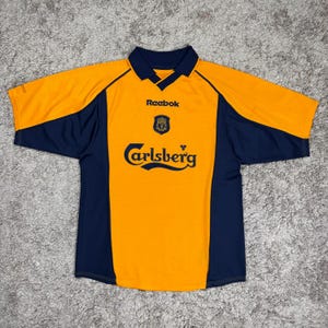 May include: An orange and navy blue short-sleeved soccer jersey. The jersey features the Reebok logo, a team crest, and the Carlsberg logo. The jersey has a collared neckline and is made of a breathable material.