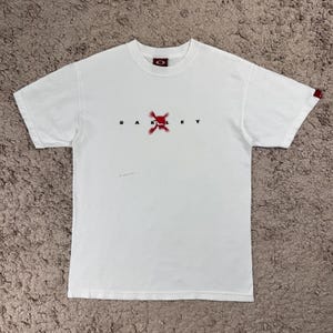 May include: White t-shirt with the brand name "OAKLEY" printed in black, with a red graphic in the center. The shirt has a small red logo near the collar and on the sleeve.