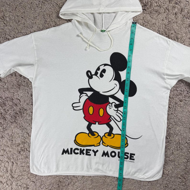 Może przedstawiać: Biała bluza z kapturem z dużą grafiką Myszki Miki w kolorze czarnym, czerwonym, ż&oacute;łtym i białym. Tekst "MICKEY MOUSE" jest wydrukowany pod postacią. Bluza jest mierzona taśmą mierniczą.