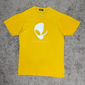 May include: Yellow t-shirt with a white alien head graphic and the text "Coming Soon". The short-sleeved shirt is made of a soft material.