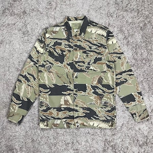 May include: A button-up jacket with a camouflage pattern in shades of green, brown, black, and white. The jacket has two chest pockets and a collar. The fabric features a leaf-like design.