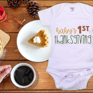 Puede incluir: Un body blanco para bebé con el texto "baby's 1st thanksgiving" en letras marrones y verdes. En el fondo, hay una porción de pastel de calabaza con crema batida en un plato blanco.