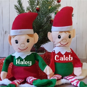 May include: Two plush elf dolls with red hats, green and red outfits, and white collars. One doll has "Haley" printed on the chest, the other has "Caleb". The dolls are sitting on a red and white surface, with a small Christmas tree in the background.