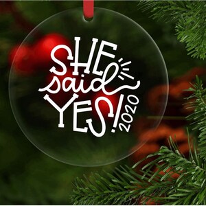 May include: Clear round Christmas ornament with the text "SHE said YES! 2020" in white lettering.