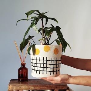 May include: A white plant pot with a black grid pattern and yellow polka dots, holding a green plant. A small brown glass bottle with reeds is next to the pot. The pot is being held by a person.