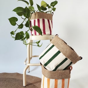 May include: Three canvas plant pots with vertical stripes in red, green, and orange. Each pot has a burlap top and leather handles. One pot holds a green leafy plant. The pots are stacked on a neutral background.