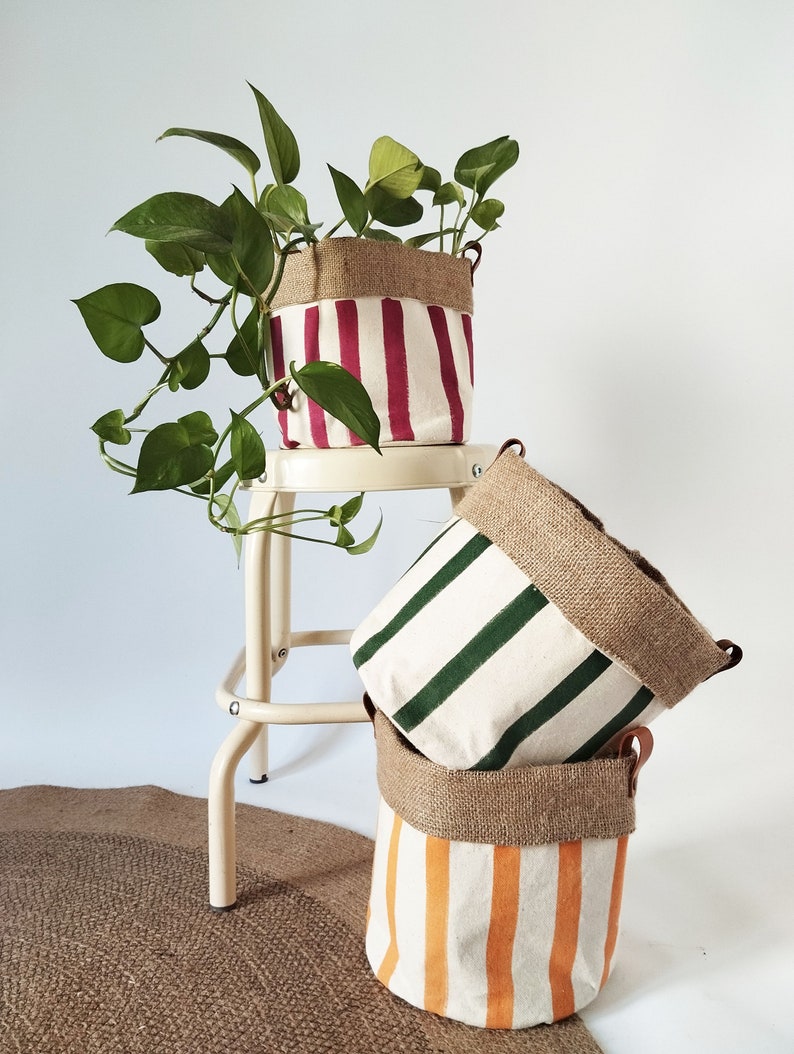 May include: Three canvas plant pots with vertical stripes in red, green, and orange. Each pot has a burlap top and leather handles. One pot holds a green leafy plant. The pots are stacked on a neutral background.