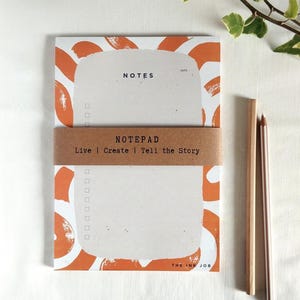 Puede incluir: Bloc de notas naranja y blanco con el texto "NOTES" y "NOTEPAD" en la parte delantera. El bloc de notas tiene una banda de papel marrón con el texto "Live | Create | Tell the Story" en él. El bloc de notas está sobre una superficie blanca con dos lápices de madera al lado.