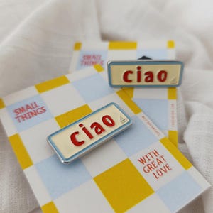 Peut inclure: Épinglette émaillée avec le mot "ciao" en rouge sur un fond crème, encadré d'un liseré bleu clair. L'épinglette est présentée sur une carte à motif quadrillé jaune et bleu, avec les mots "Small Things" et "With Great Love".