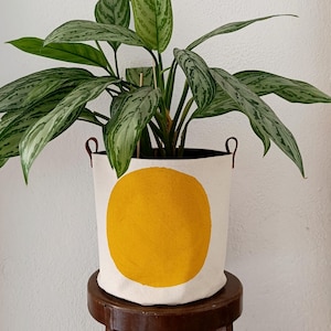 Può includere: Un coprivaso cilindrico in tessuto color bianco sporco con un grande cerchio giallo pieno dipinto sul davanti. Il contenitore presenta due piccole maniglie laterali in similpelle marrone e contiene una pianta d'appartamento con foglie verdi e argentee.