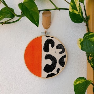 Op de afbeelding: Een ronde stoffen wandhanger in een houten borduurring. Het ontwerp heeft een feloranje gedeelte aan de linkerkant en een crèmekleurig gedeelte met zwarte abstracte vormen aan de rechterkant. De ring wordt opgehangen met een houten peg en een leren koord.