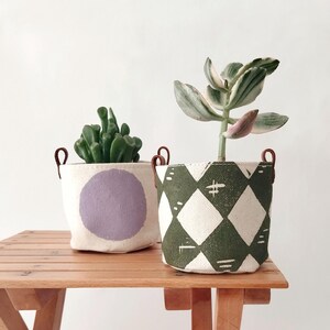 以下が含まれることがあります： 木製のテーブルに、革の持ち手が付いた2つの布製植木鉢があります。1つはラベンダー色の円が付いたクリーム色で、もう1つはひし形模様の緑と白です。どちらの鉢にも植物が入っています。