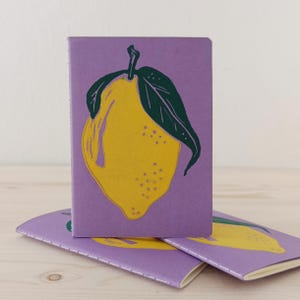 May include: A set of three rectangular notebooks with a light purple cover. Each notebook features a central graphic of a bright yellow lemon with a dark green leaf. The notebooks are bound with visible white stitching along the spine edge.