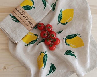 Puede incluir: Un paño de cocina de lino beige con un patrón repetitivo de limones amarillos y hojas verde oscuro. Una sección doblada está envuelta en una banda de papel con el texto 100% linen, PRINTED BY HAND y take care of me. Un racimo de tomates cherry rojos.