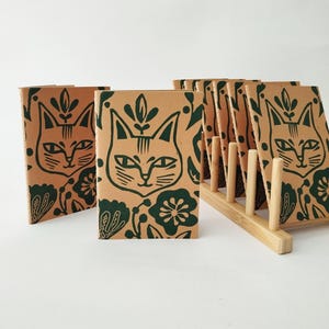 May include: A collection of tan paper notebooks featuring a dark green screen-printed cat face and floral pattern. One notebook stands alone in the foreground, while several others are organized in a light-colored wooden rack stand.