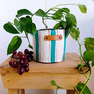 Puede incluir: Una maceta cilíndrica de tela con rayas verticales de color verde azulado y finas rayas rojas sobre fondo blanco, con un asa de cuero color canela sujeta por dos remaches metálicos color latón, que contiene una planta verde con enredaderas.