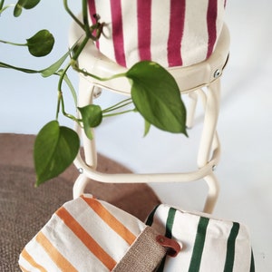 May include: Three canvas plant pots with vertical stripes in orange, green, and burgundy. The pots have a burlap trim and are sitting on a white stool and a brown rug. A green plant with heart-shaped leaves is in the background.