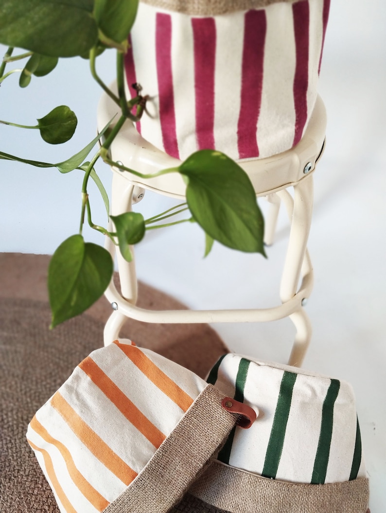 May include: Three canvas plant pots with vertical stripes in orange, green, and burgundy. The pots have a burlap trim and are sitting on a white stool and a brown rug. A green plant with heart-shaped leaves is in the background.