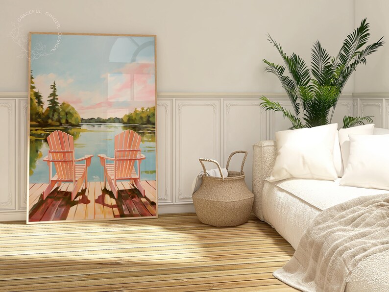 May include: A framed art print depicts two orange Adirondack chairs on a wooden dock overlooking a lake. The artwork features a serene landscape with trees, water, and a colorful sky. The scene evokes a sense of relaxation and leisure.
