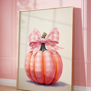 A framed painting of a pumpkin adorned with a large pink and white checkered bow. The pumpkin has a soft, pastel, pink and white check pattern and is set against a light background. The frame leans against a pink paneled wall on a wooden floor.
