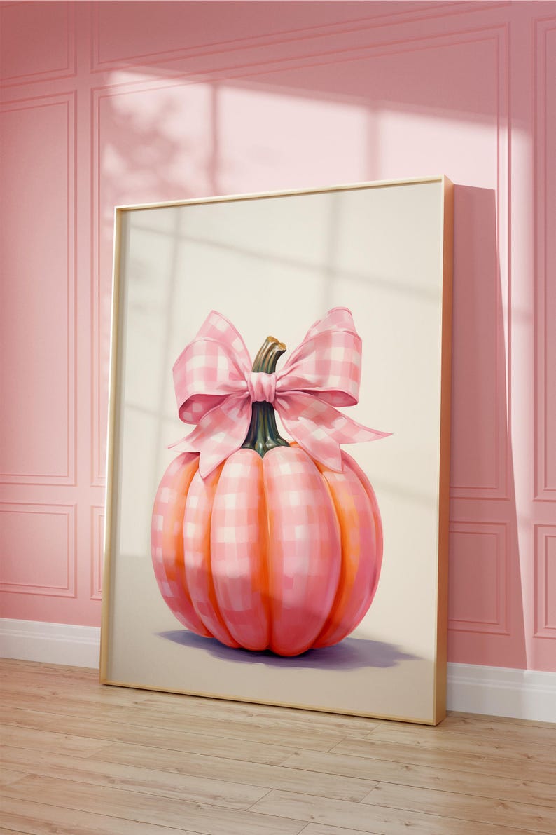 A framed painting of a pumpkin adorned with a large pink and white checkered bow. The pumpkin has a soft, pastel, pink and white check pattern and is set against a light background. The frame leans against a pink paneled wall on a wooden floor.