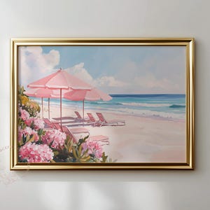 Framed coastal artwork featuring pink umbrellas and lounge chairs on a pastel shoreline. The piece hangs on a neutral-toned wall, accented by golden framing and soft natural light, creating a relaxed beach house atmosphere.
