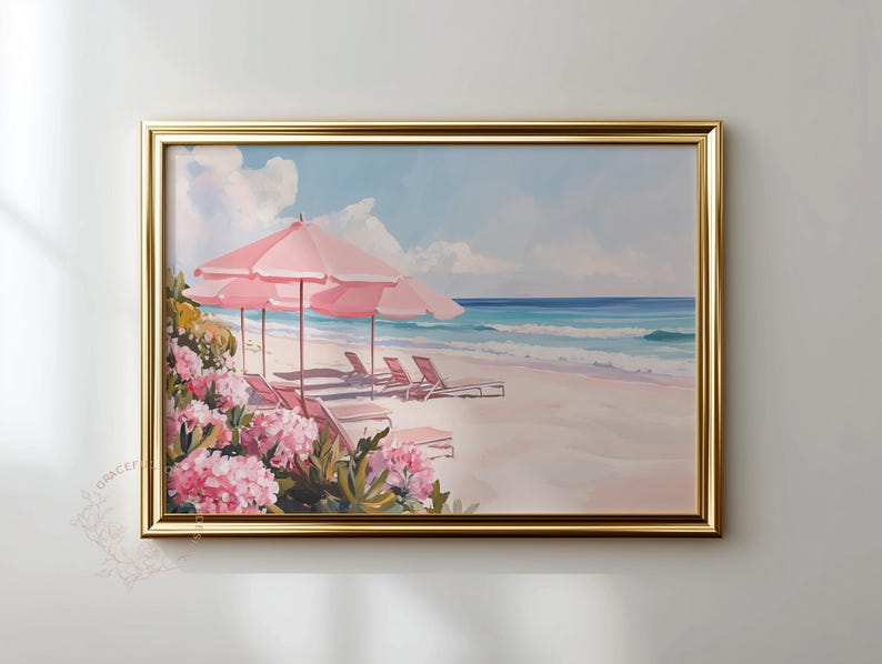 Framed coastal artwork featuring pink umbrellas and lounge chairs on a pastel shoreline. The piece hangs on a neutral-toned wall, accented by golden framing and soft natural light, creating a relaxed beach house atmosphere.