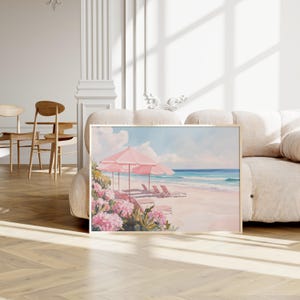 Framed beach painting resting against a plush cream sofa in a bright, elegant room. The artwork features soft pink beach umbrellas, lounge chairs, and blooming coastal flowers set along a sunlit shoreline with calm waves.