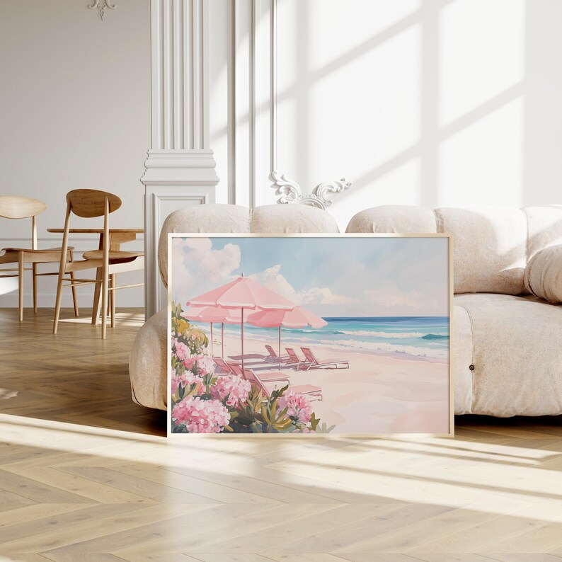 Framed beach painting resting against a plush cream sofa in a bright, elegant room. The artwork features soft pink beach umbrellas, lounge chairs, and blooming coastal flowers set along a sunlit shoreline with calm waves.