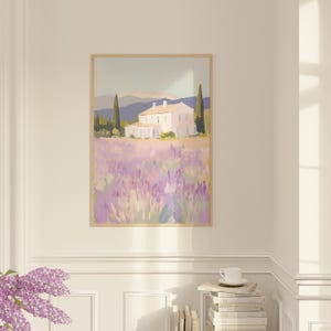Framed lavender field painting hung in a bright corner above a stack of books and a cup. The peaceful countryside scene brings warmth and calm to the refined white-paneled room.