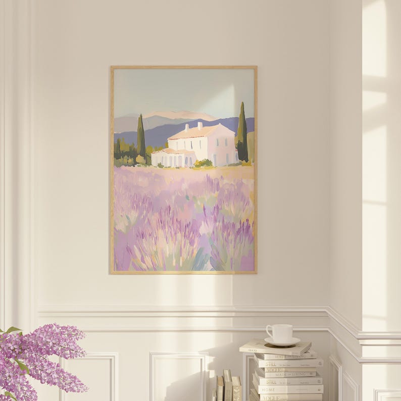 Framed lavender field painting hung in a bright corner above a stack of books and a cup. The peaceful countryside scene brings warmth and calm to the refined white-paneled room.