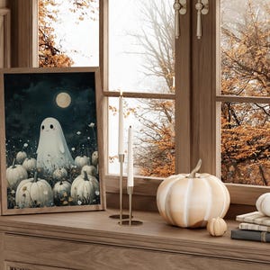 May include: A framed art print featuring a friendly ghost in a field of pumpkins under a full moon. A large white pumpkin, small pumpkins, and candles sit on a wooden surface near a window with fall foliage.
