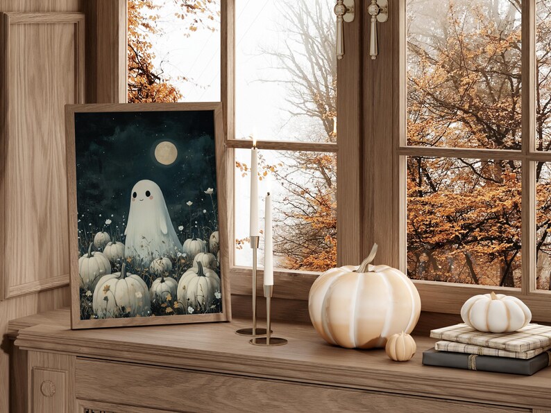 May include: A framed art print featuring a friendly ghost in a field of pumpkins under a full moon. A large white pumpkin, small pumpkins, and candles sit on a wooden surface near a window with fall foliage.