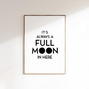 May include: A framed print with the text "IT'S ALWAYS A FULL MOON IN HERE" in black against a white background. The frame is a light brown color, and the print is hanging on a white wall.