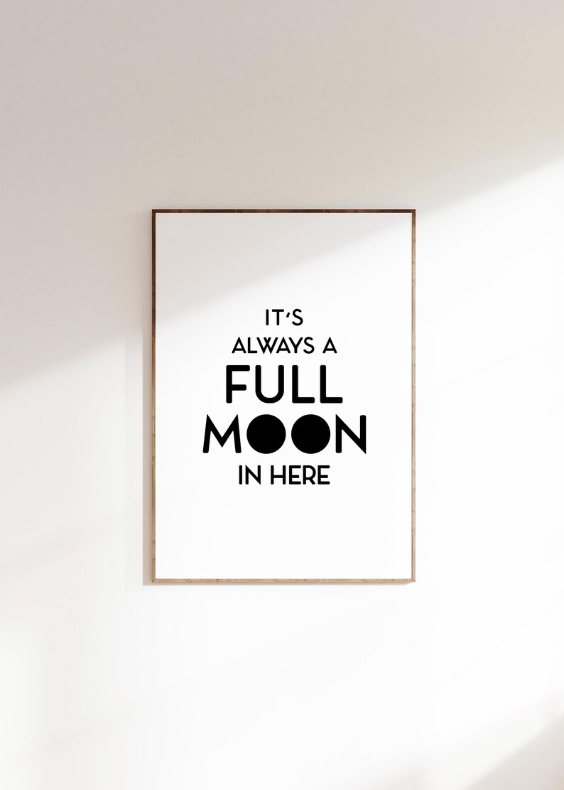 May include: A framed print with the text "IT'S ALWAYS A FULL MOON IN HERE" in black against a white background. The frame is a light brown color, and the print is hanging on a white wall.