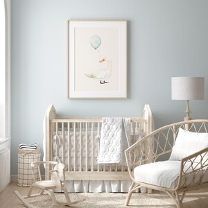 May include: A framed watercolor print of a white duck holding a blue balloon. The artwork hangs above a light-colored wooden crib in a nursery setting. The room has light blue walls and a wicker chair.