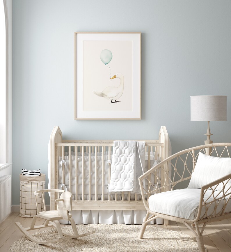 May include: A framed watercolor print of a white duck holding a blue balloon. The artwork hangs above a light-colored wooden crib in a nursery setting. The room has light blue walls and a wicker chair.