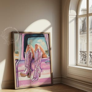 May include: Framed artwork featuring a pair of sparkly pink cowboy boots. The boots are depicted in a car window, with legs and a sky visible. The artwork is in a wooden frame and leans against a wall.