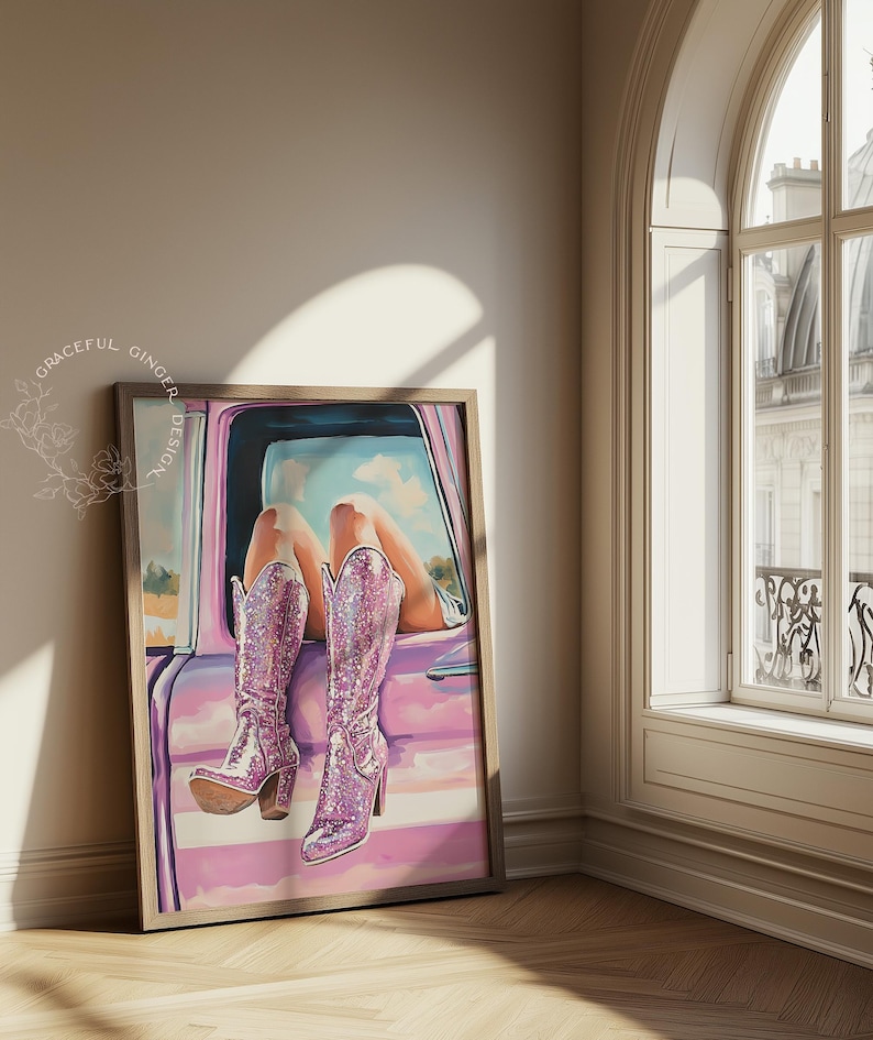 May include: Framed artwork featuring a pair of sparkly pink cowboy boots. The boots are depicted in a car window, with legs and a sky visible. The artwork is in a wooden frame and leans against a wall.