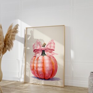 Puede incluir: Impresi&oacute;n enmarcada de una calabaza rosa adornada con un gran lazo de cuadros rosa y blanco. La calabaza muestra tonos de rosa y naranja, con un tallo verde. La obra de arte est&aacute; sobre un fondo neutro, creando un encantador tema oto&ntilde;al.
