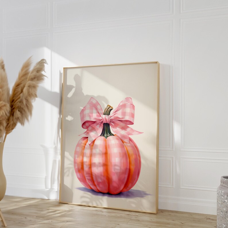 Puede incluir: Impresi&oacute;n enmarcada de una calabaza rosa adornada con un gran lazo de cuadros rosa y blanco. La calabaza muestra tonos de rosa y naranja, con un tallo verde. La obra de arte est&aacute; sobre un fondo neutro, creando un encantador tema oto&ntilde;al.