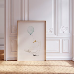 May include: A framed watercolor print featuring a white goose holding a light blue balloon. The goose has a yellow beak and black feet. The artwork is displayed in a room with white paneling and a wooden floor, creating a soft, whimsical aesthetic.