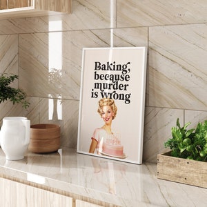 Peut inclure: Une impression encadr&eacute;e blanche avec le texte "Baking, because murder is wrong." Une femme aux cheveux blonds tient un g&acirc;teau rose avec des bougies.